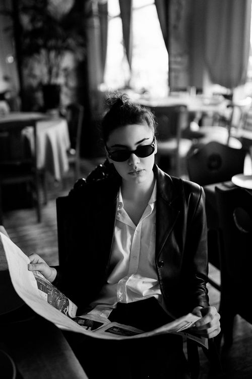 Free A fashionable woman in sunglasses reads a newspaper inside a cozy café, captured in black and white. Stock Photo