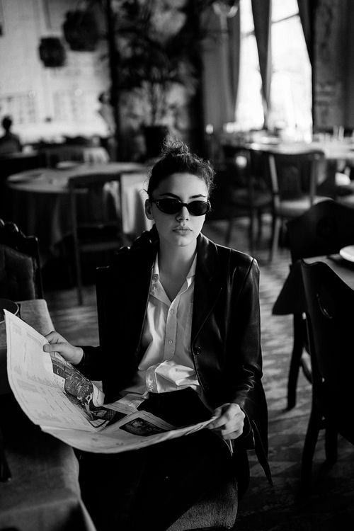 Free Elegant black and white portrait of a woman in sunglasses reading a newspaper in a chic café. Stock Photo