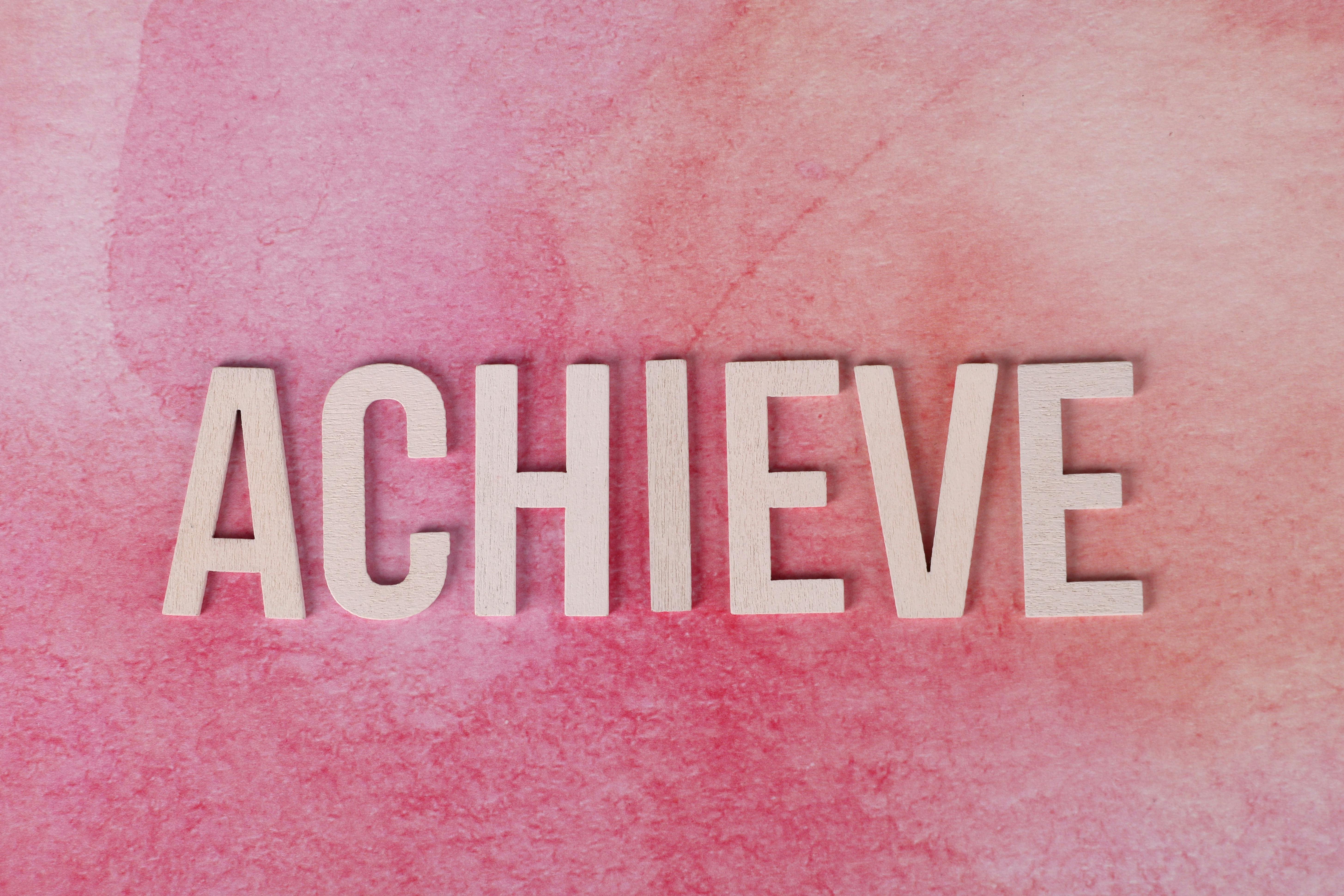 The word 'Achieve' in wooden letters on a textured pink background, symbolizing success and motivation.