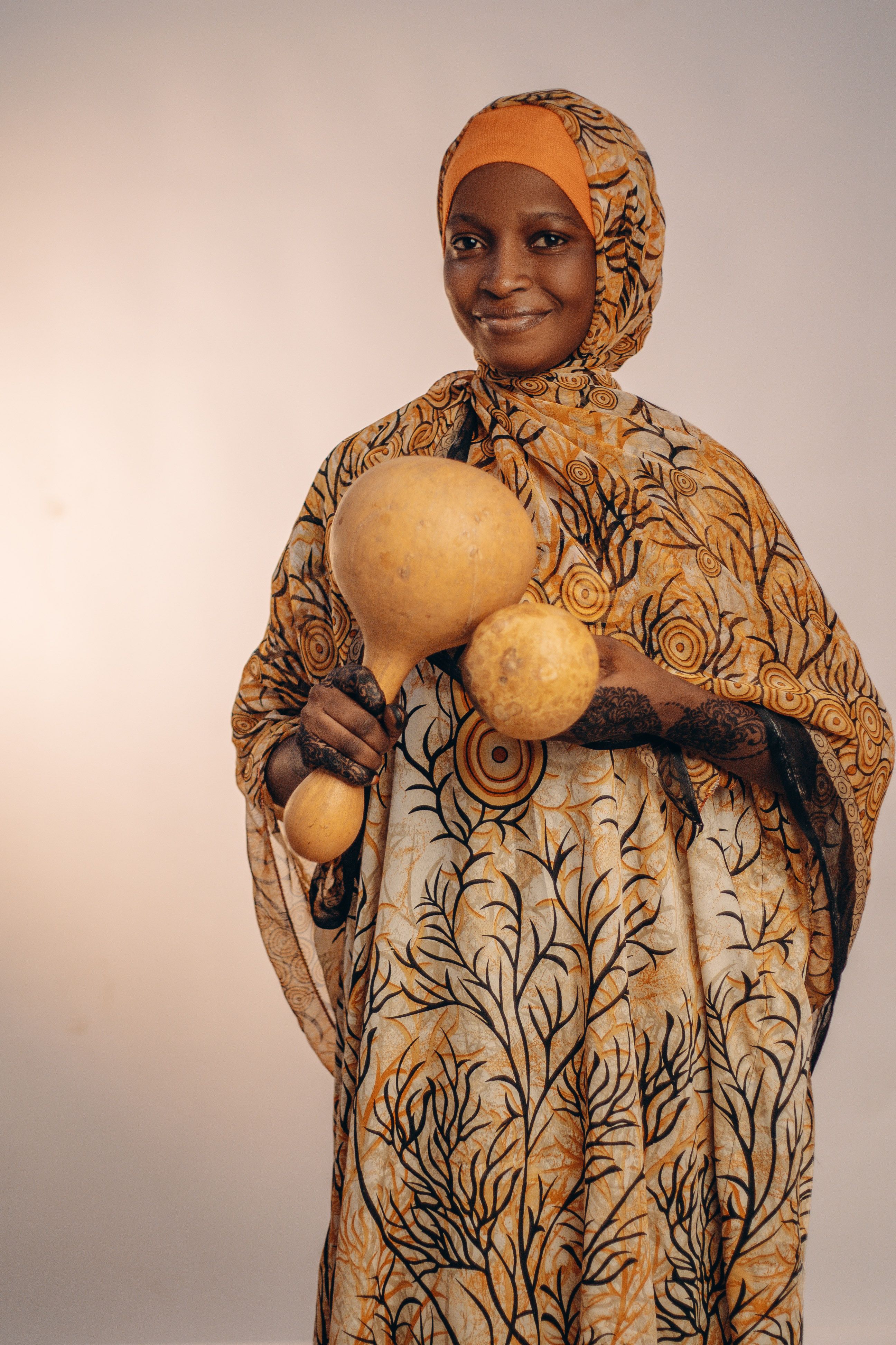 Smiling African woman in traditional attire holding three calabashes against a neutral background.