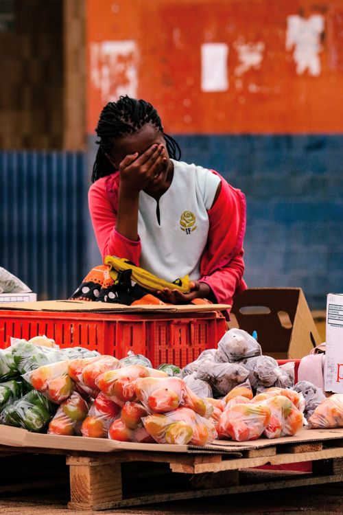 Free A young vendor in Nhlangano, Eswatini, sitting behind a colorful array of produce at an outdoor market. Stock Photo
