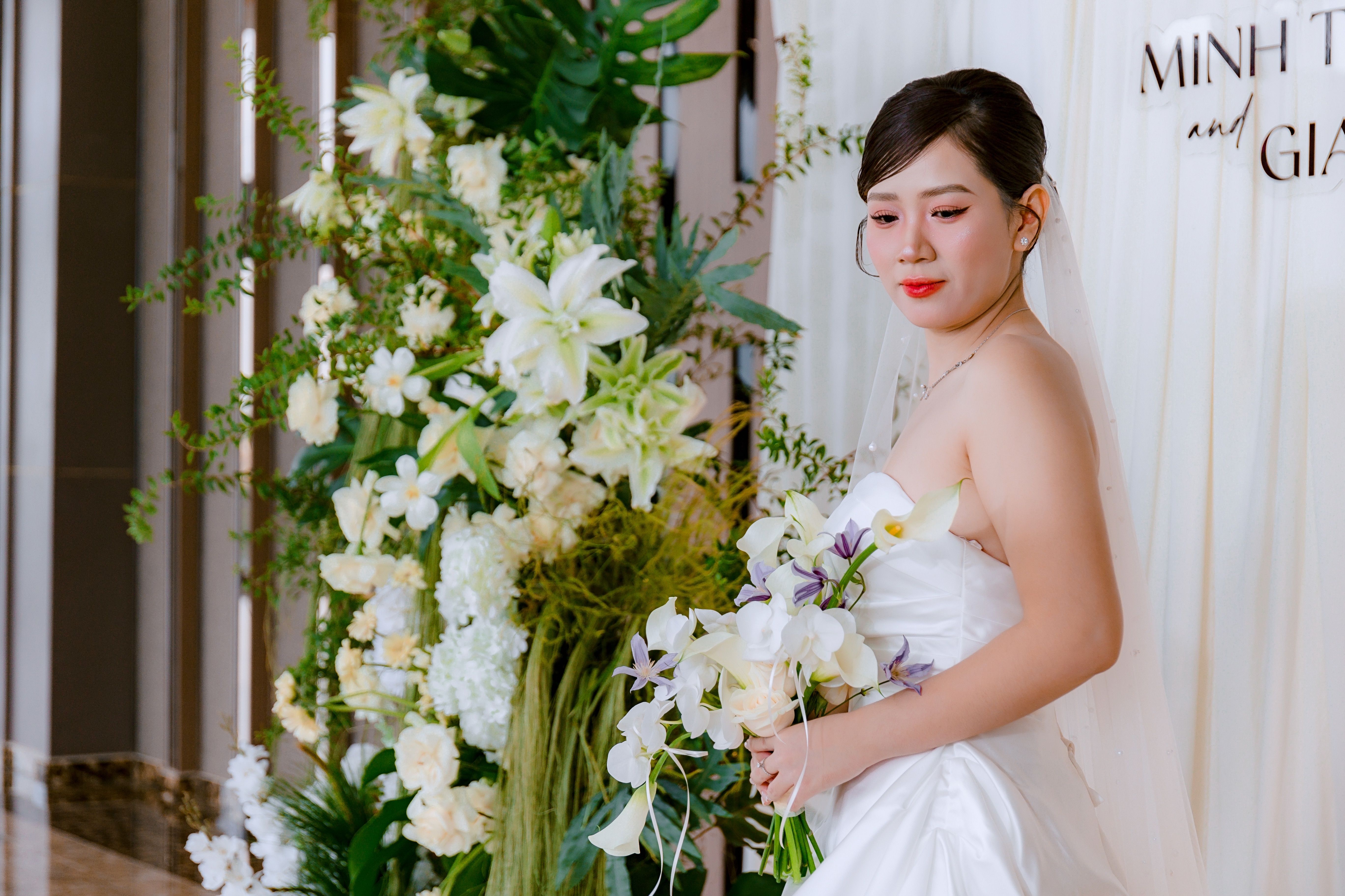 Bride in white dress with floral arrangement indoors, holding bouquet.