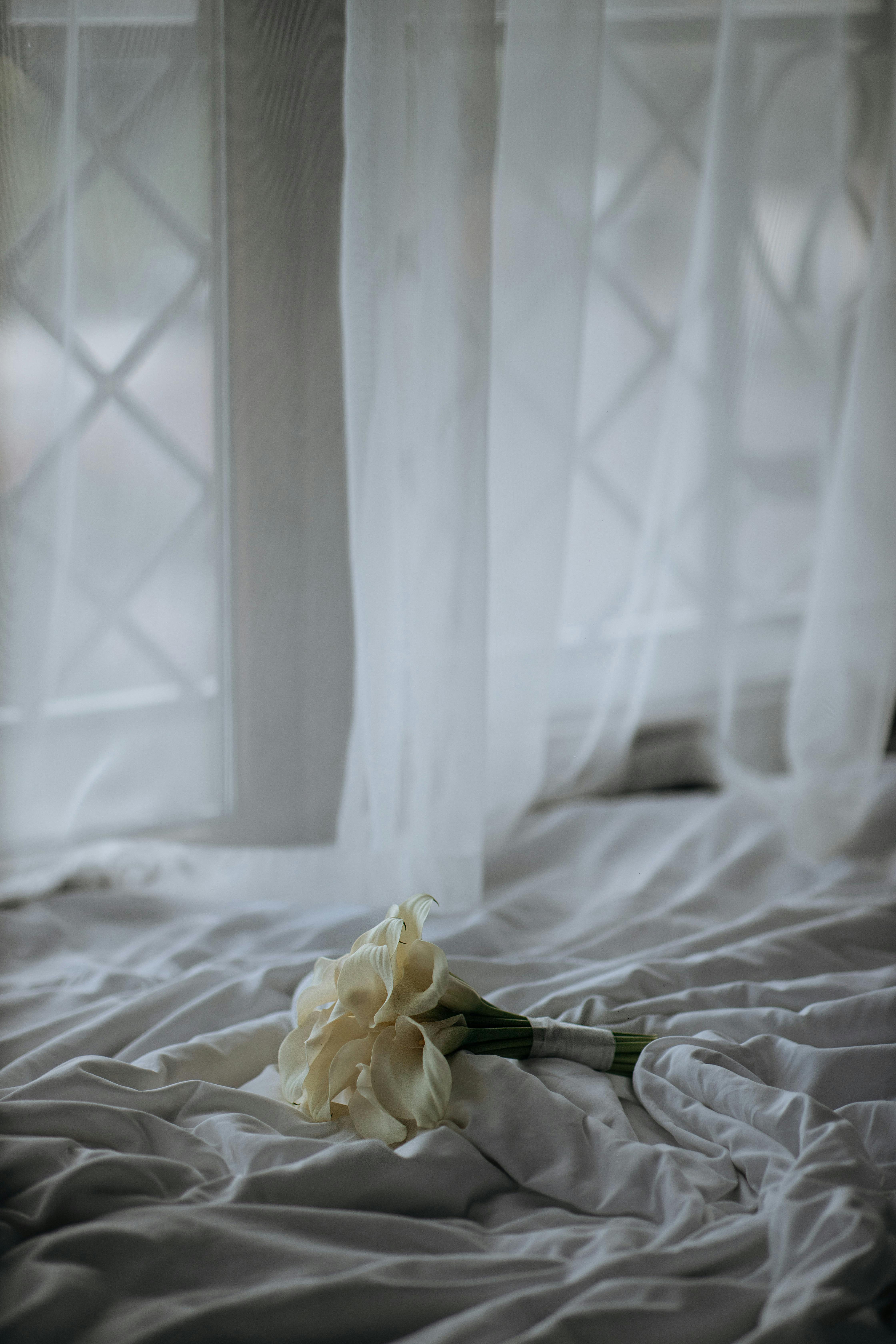 A beautiful white calla lily bouquet rests on a soft, draped fabric beside a window with sheer curtains.
