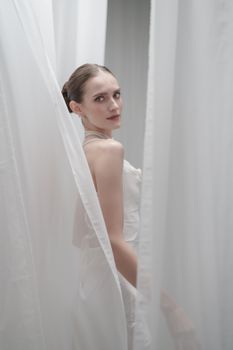 Side view of a woman in a white dress standing elegantly between sheer white curtains.