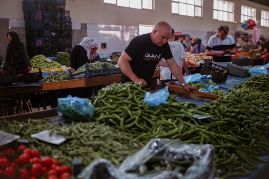 Indoor market scene with people shopping for fresh green peppers and various produce.