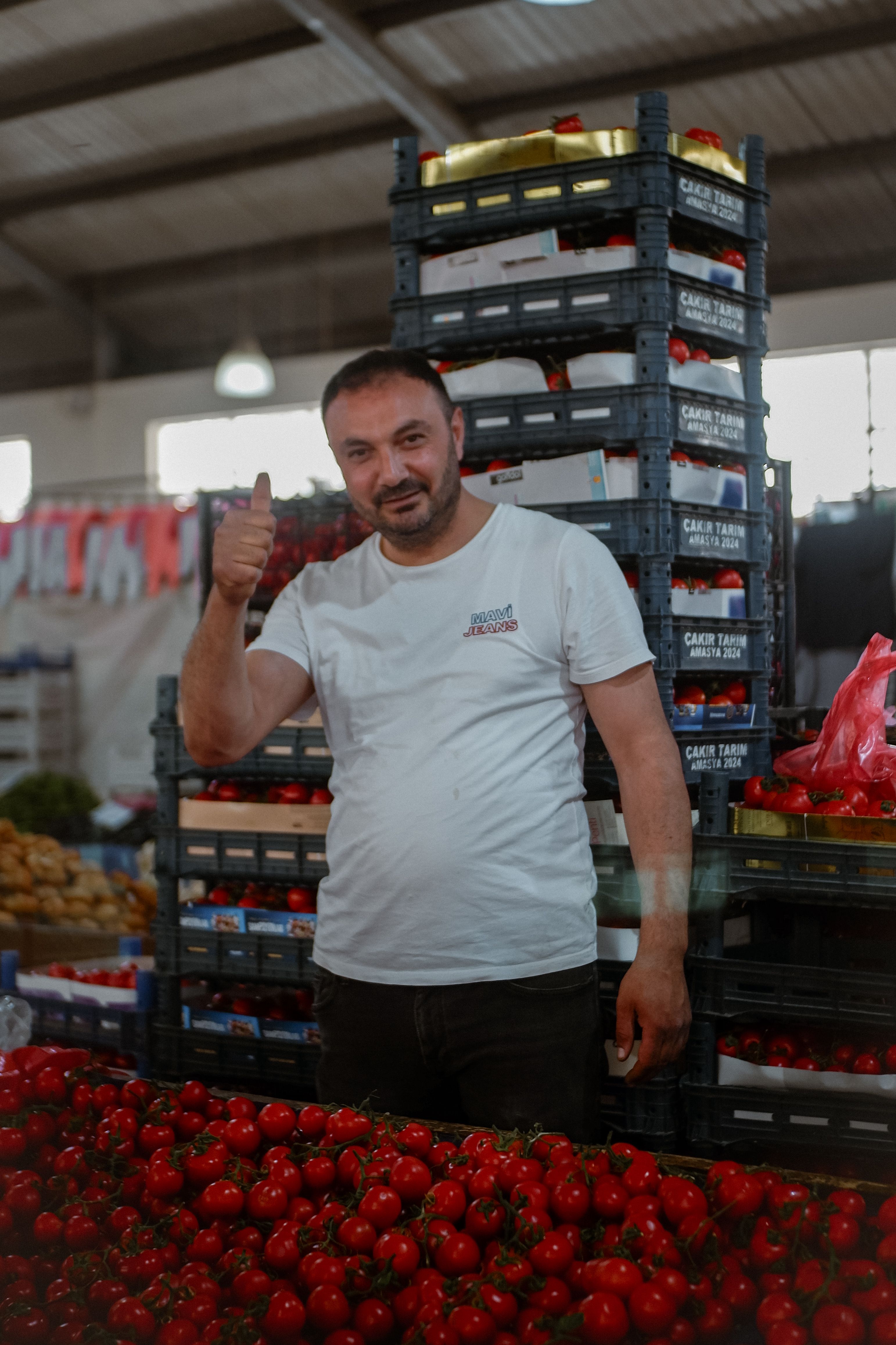 Friendly vendor giving thumbs up at a vibrant farmer's market stand with fresh tomatoes.