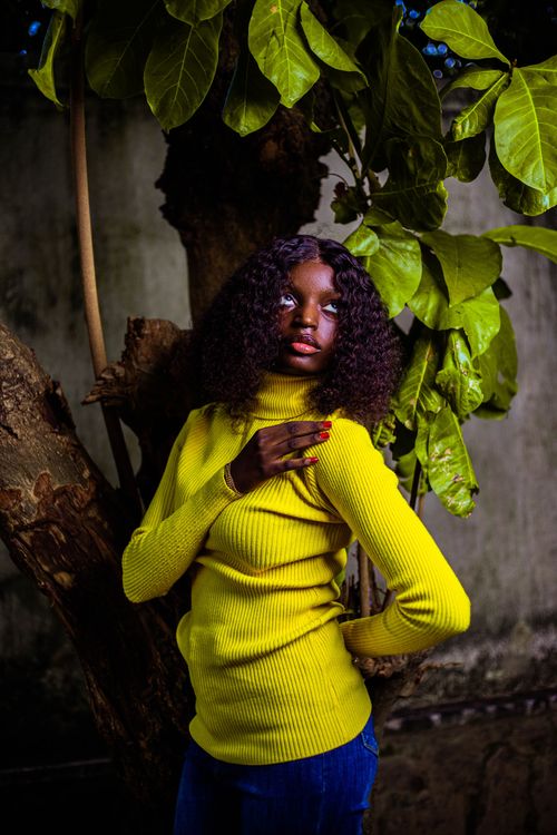 Free A striking portrait of a woman in a yellow sweater standing under vibrant green leaves. Stock Photo