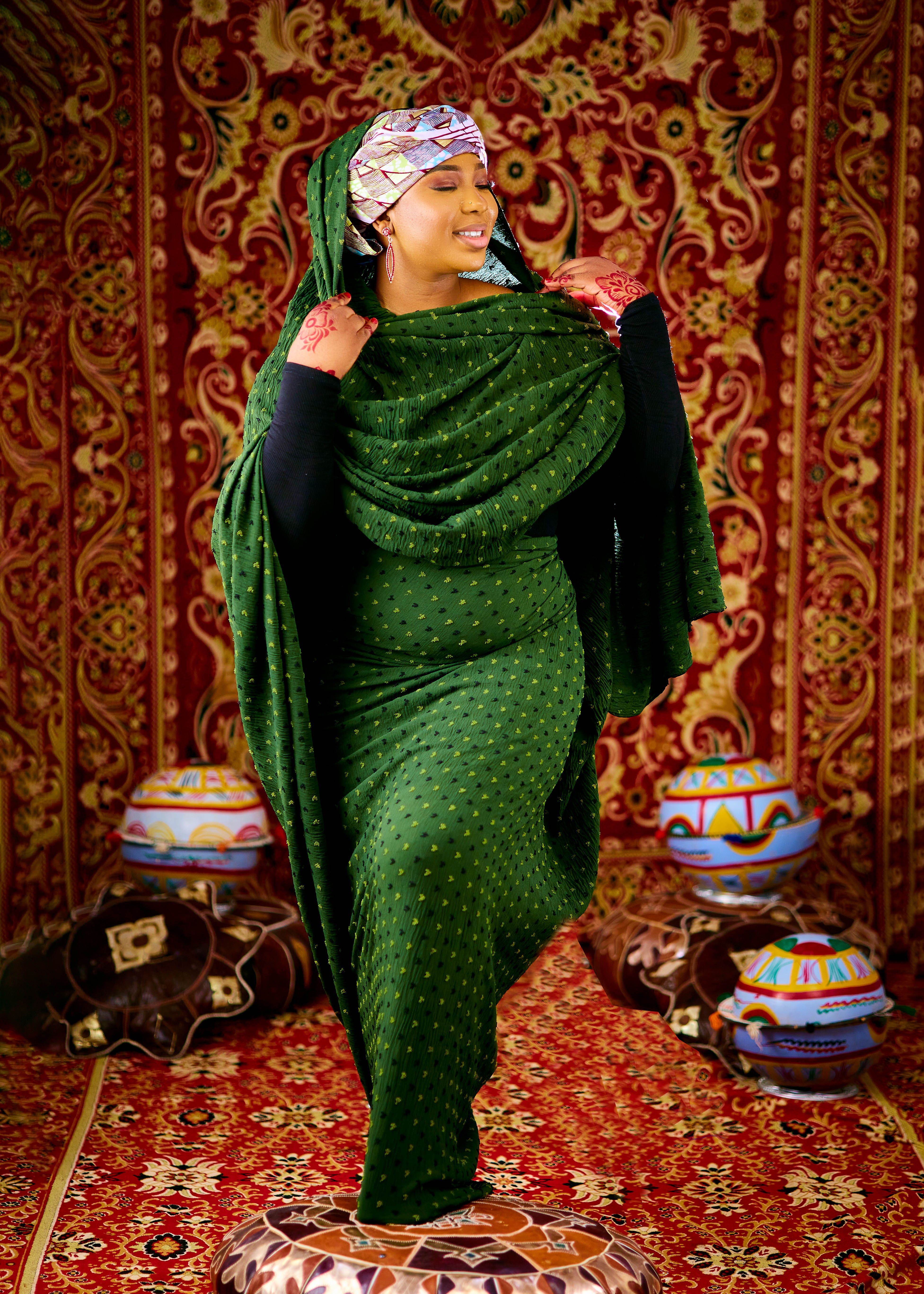 Woman in green traditional dress with intricate patterns indoors, surrounded by colorful decor.