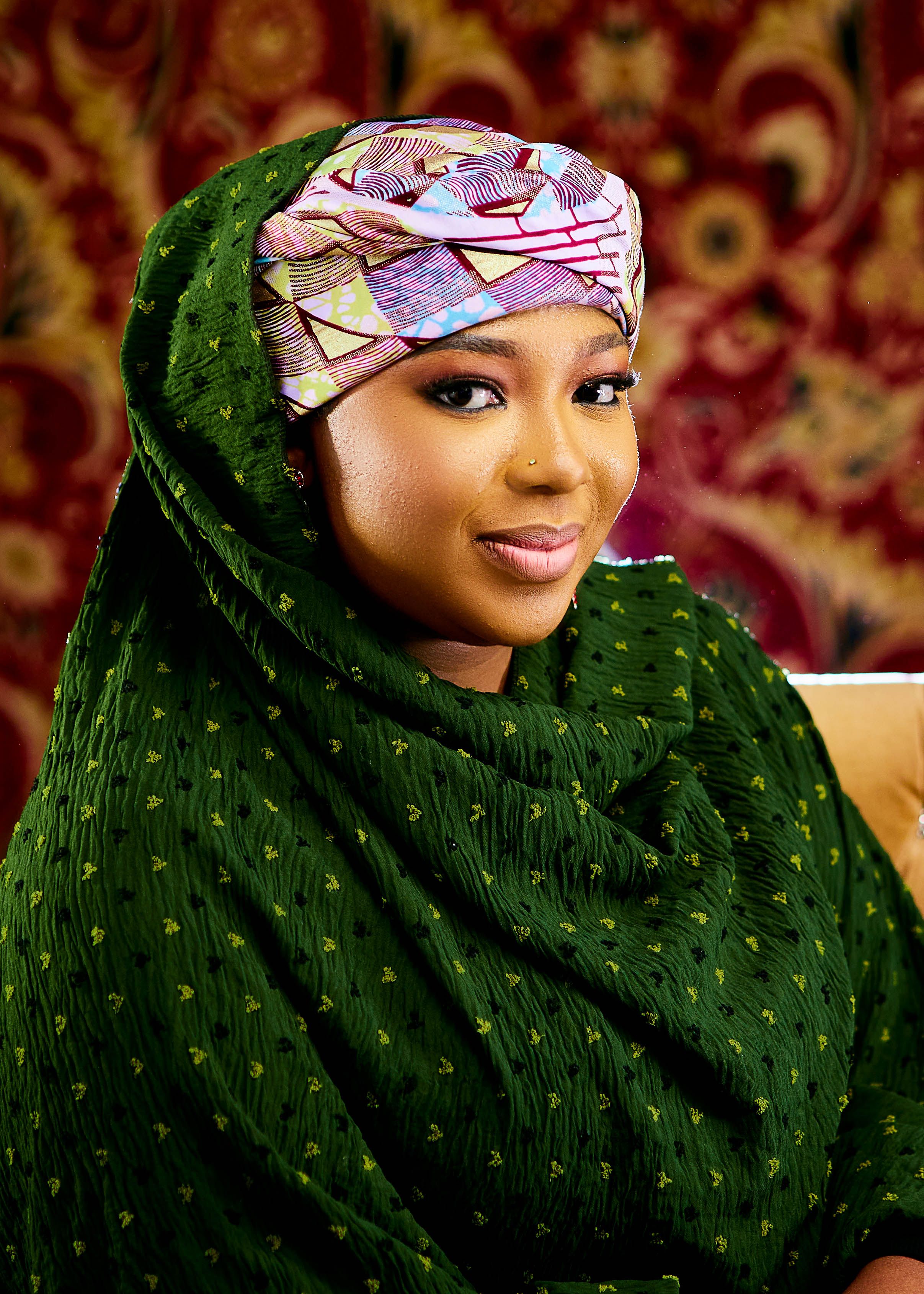 A beautiful woman in traditional green attire and colorful headscarf, smiling warmly indoors.