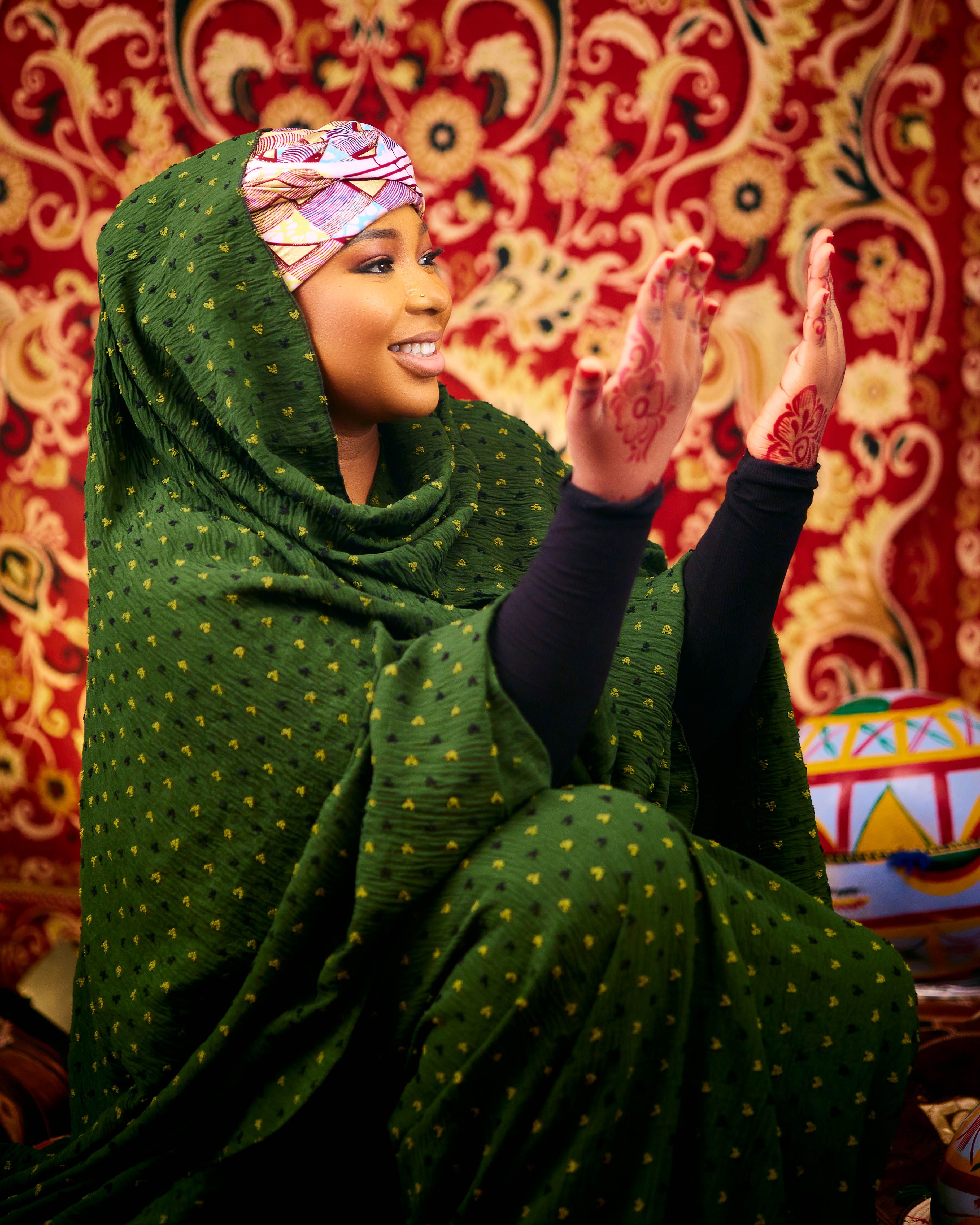 Smiling woman in green traditional dress with henna designs on hands, vibrant background.