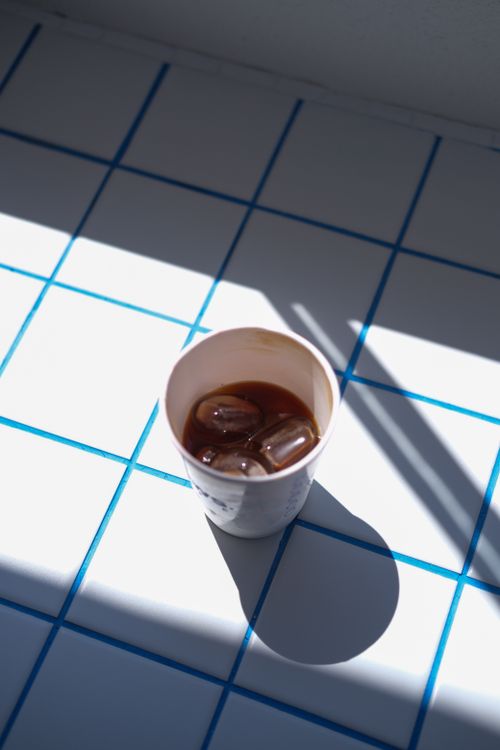 Free A cup of iced coffee casting a shadow on a blue and white tiled surface, bathed in sunlight. Stock Photo