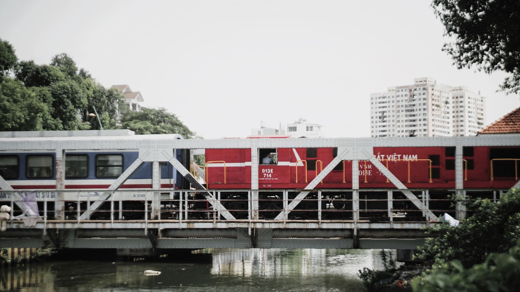Red and blue train crossing a bridge in an urban area with buildings in the background.