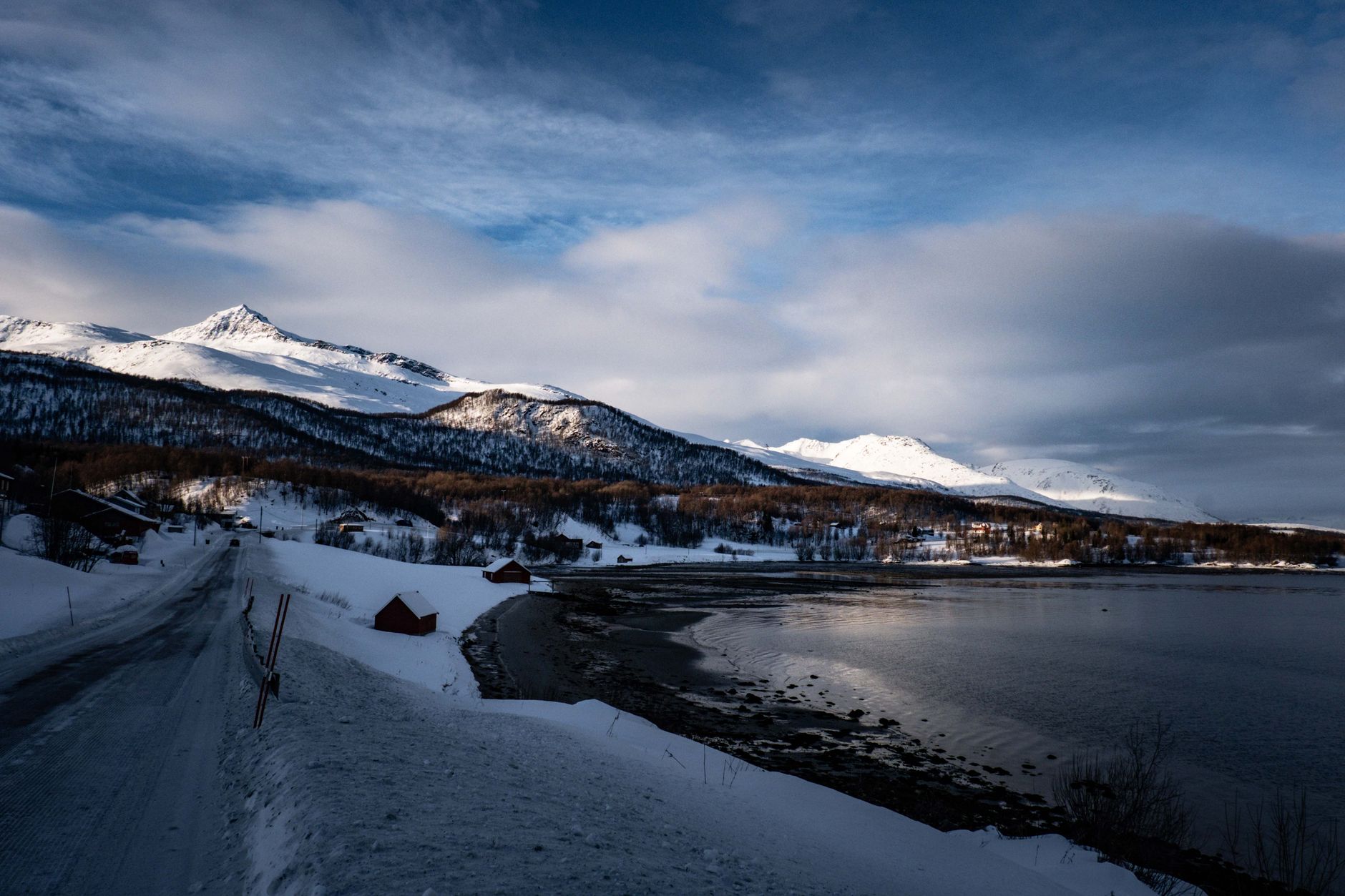 Breathtaking winter scene with snow-covered mountains and a serene waterfront under a dramatic sky.