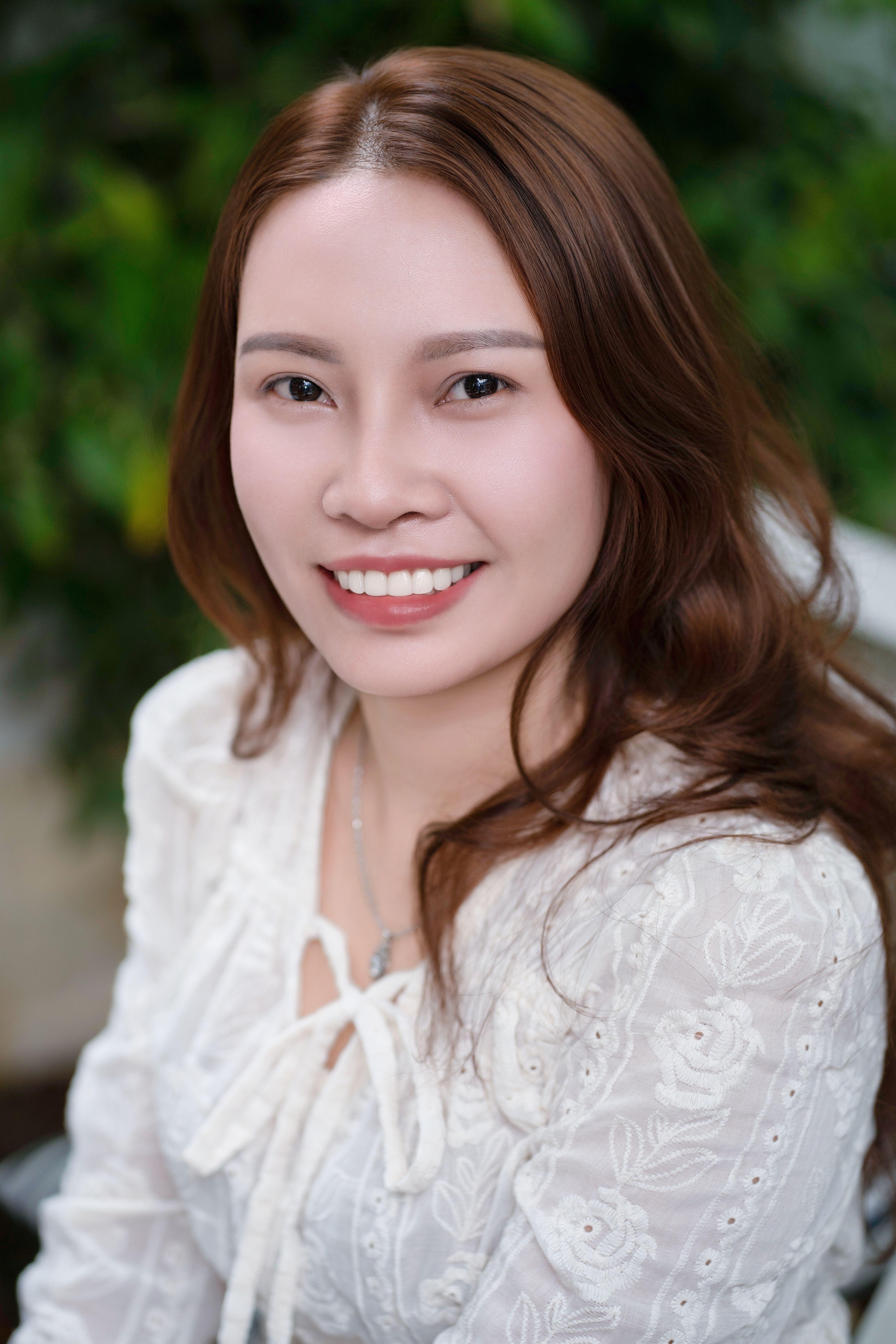 Portrait of a smiling woman in a white blouse with lush greenery background.
