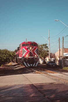 A vibrant red train travels through an urban railway crossing under a clear blue sky.