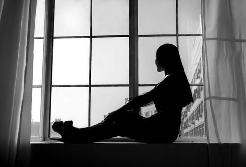 Free Black and white side view silhouette of stylish female with long hair wearing high heels sitting on windowsill and looking out Stock Photo