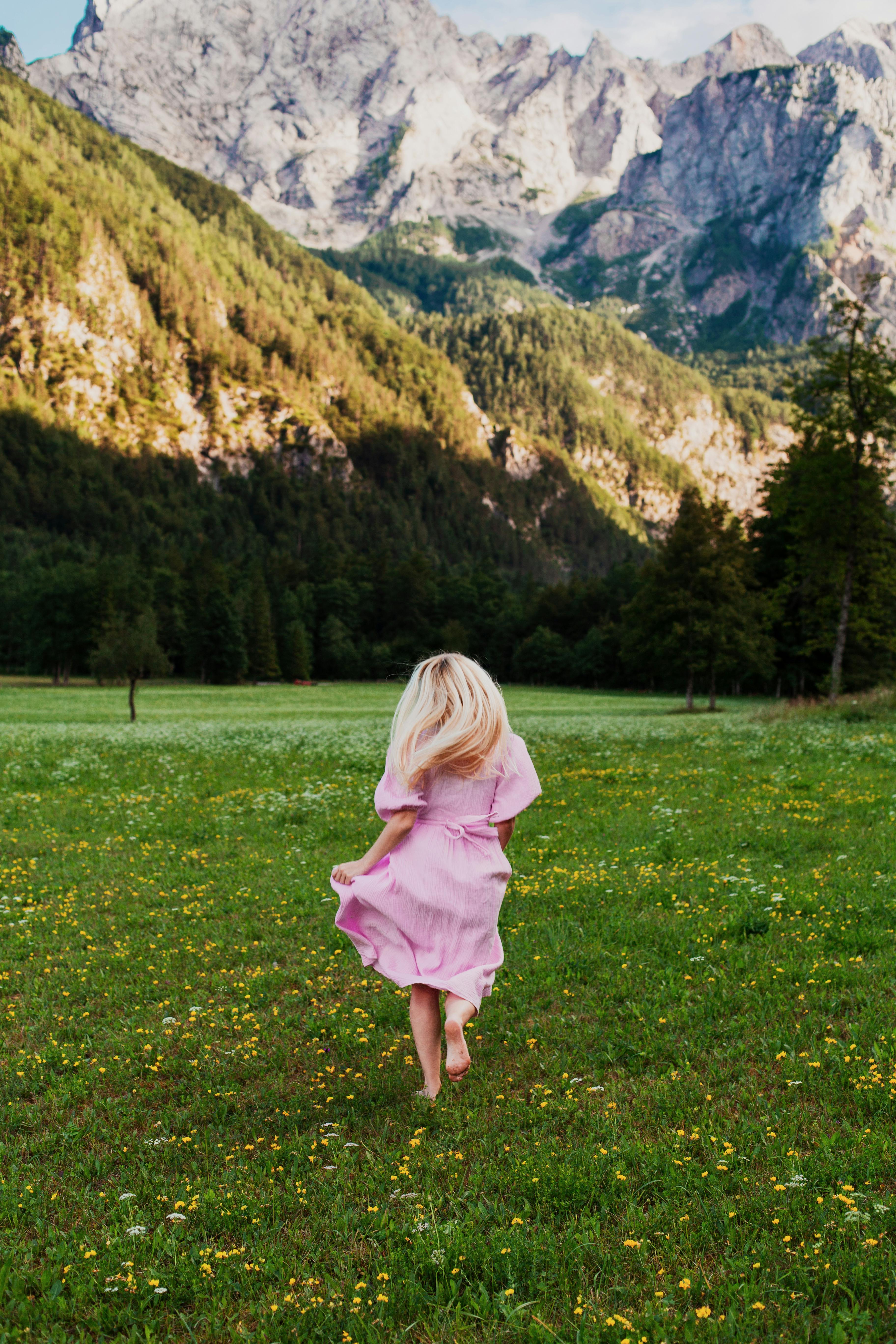 A woman in a pink dress runs through a vibrant green meadow in Slovenia with majestic mountains in the background.