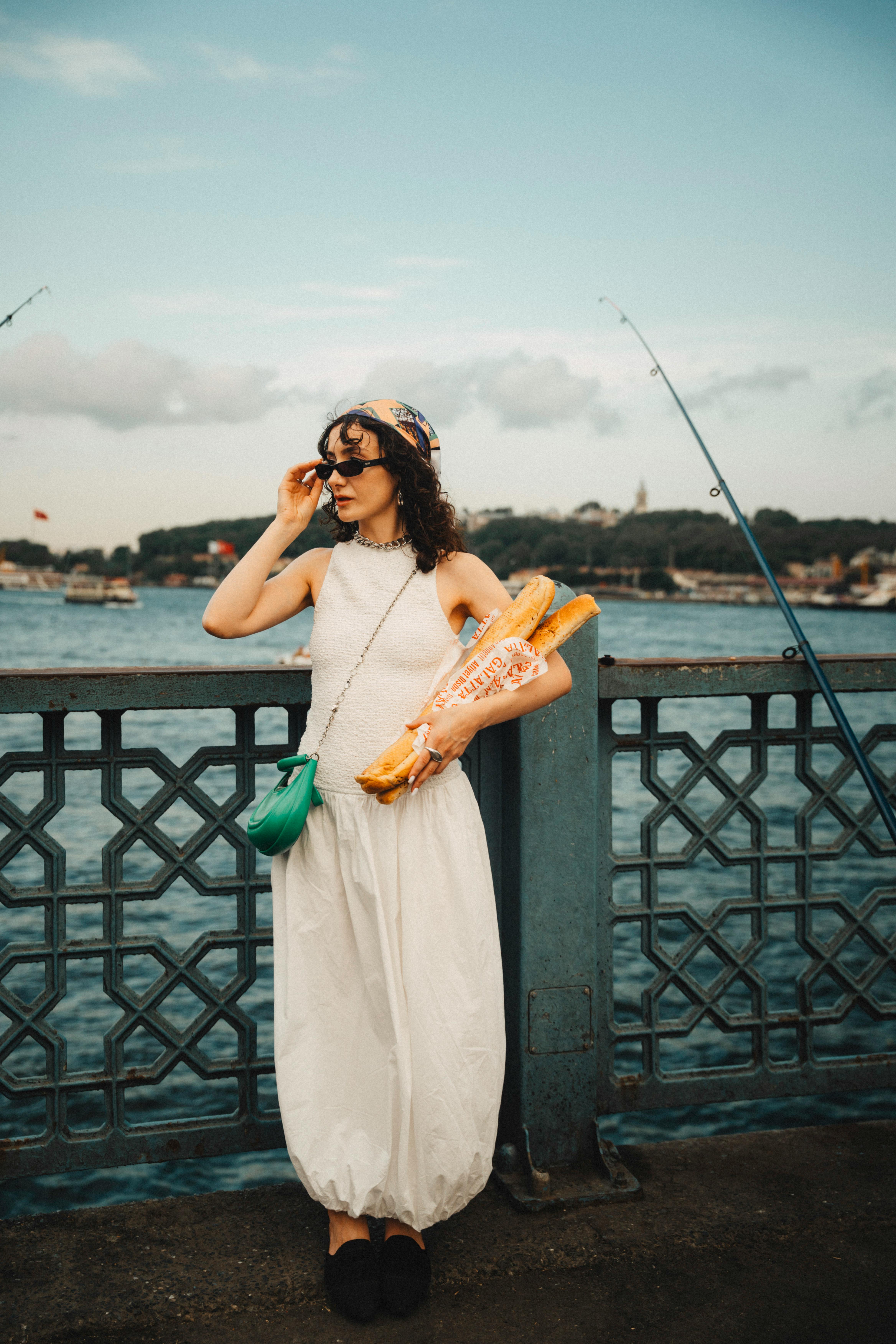 Chic woman in white dress holding baguettes on a scenic waterfront bridge.