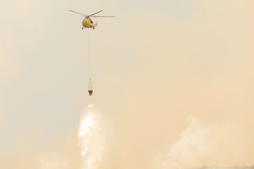 Free Yellow helicopter drops water to combat wildfires amidst smoky skies. Stock Photo