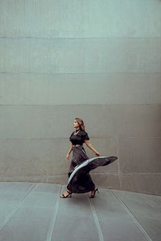 Graceful woman in a flowing black dress walks stylishly past a contemporary concrete wall.