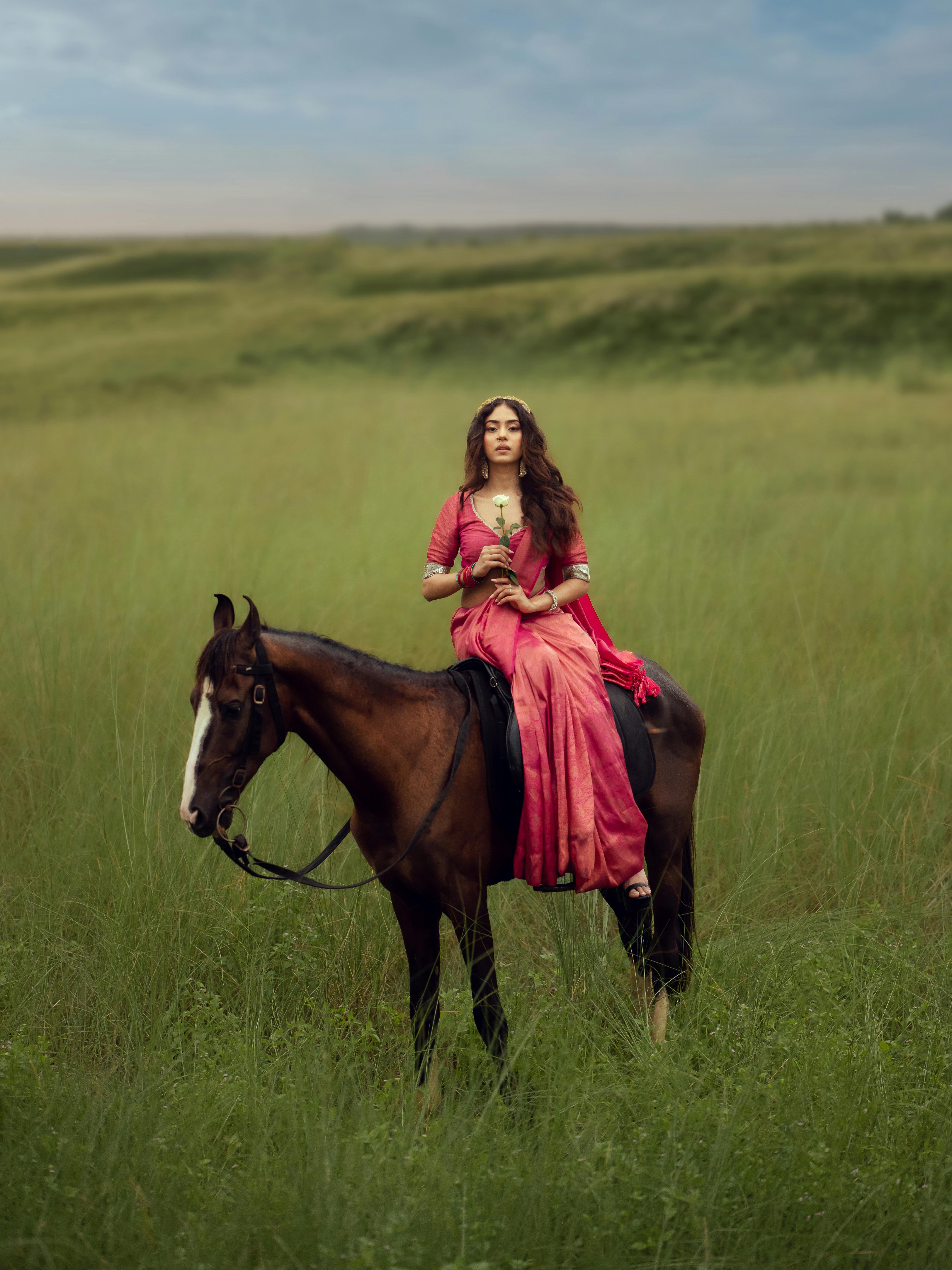 Elegant woman in red traditional attire rides a horse in lush green grassland, evoking serenity and grace.