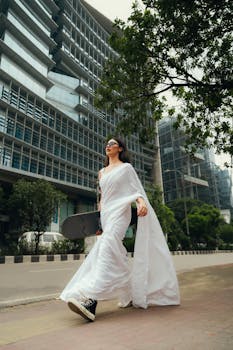 A woman in a white saree and sneakers holds a skateboard, walking through a modern urban environment.