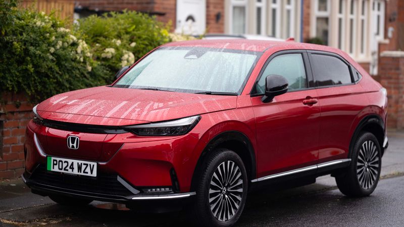An electric crossover vehicle parked in a residential driveway with a charging cable connected to a wall unit