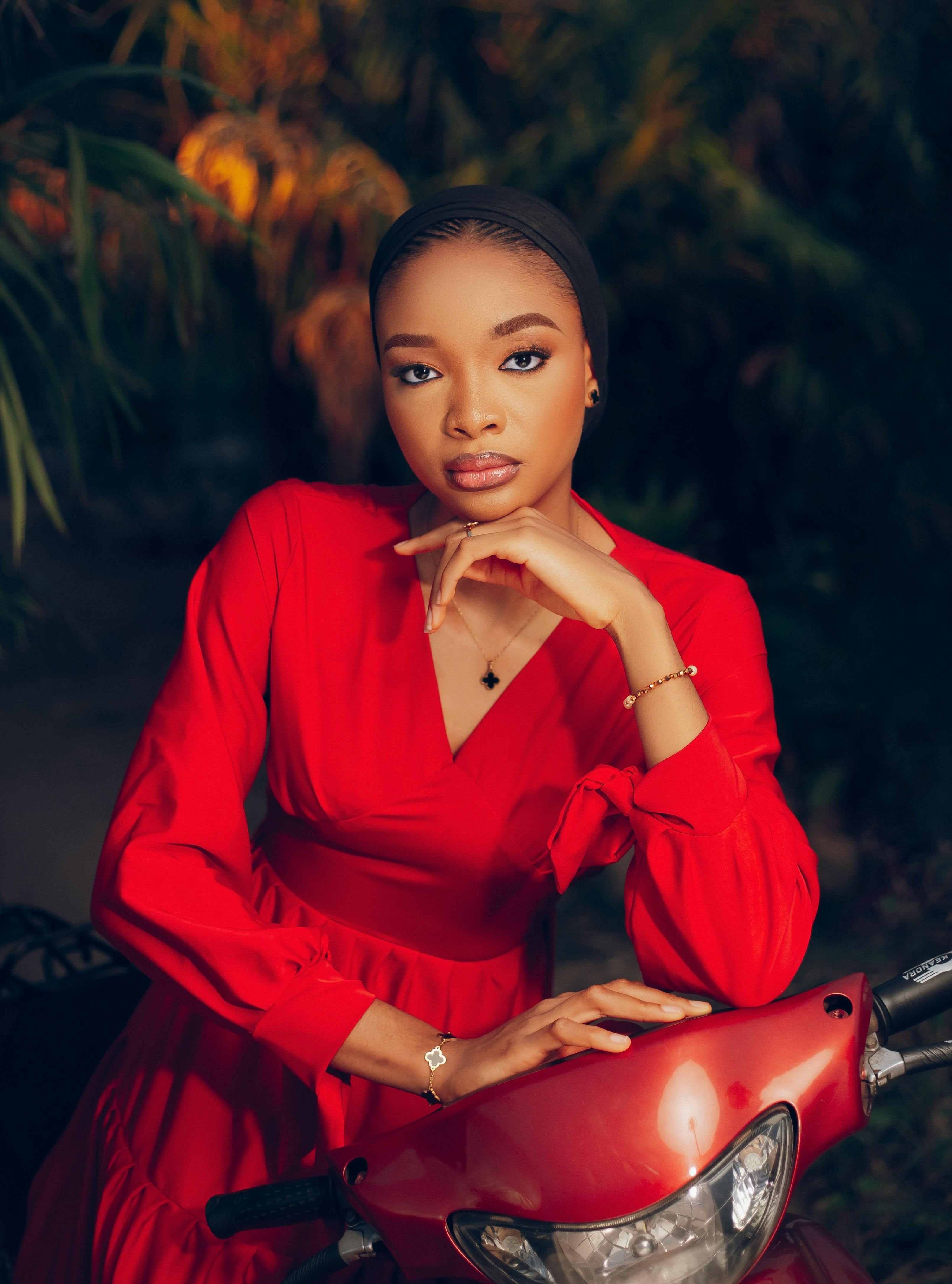 Elegant woman in red dress posing with scooter, exuding confidence outdoors.
