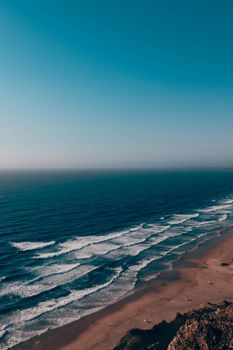 A tranquil aerial shot of ocean waves meeting a sandy beach under a clear blue sky.