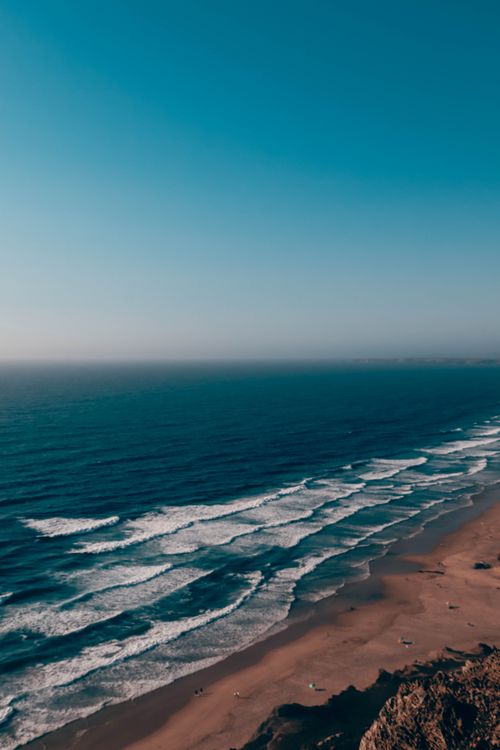 Free A tranquil aerial shot of ocean waves meeting a sandy beach under a clear blue sky. Stock Photo
