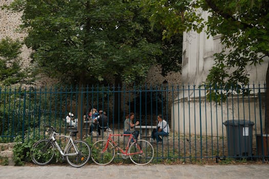 Urban park scene with people, bicycles, and greenery behind a fence.