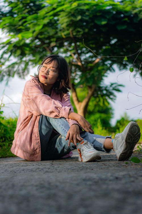 Free Young woman sitting on pavement surrounded by lush green trees under a clear blue sky. Stock Photo
