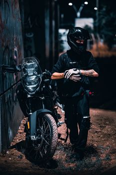 Rider in black gear standing with a motorcycle in an urban setting at night.