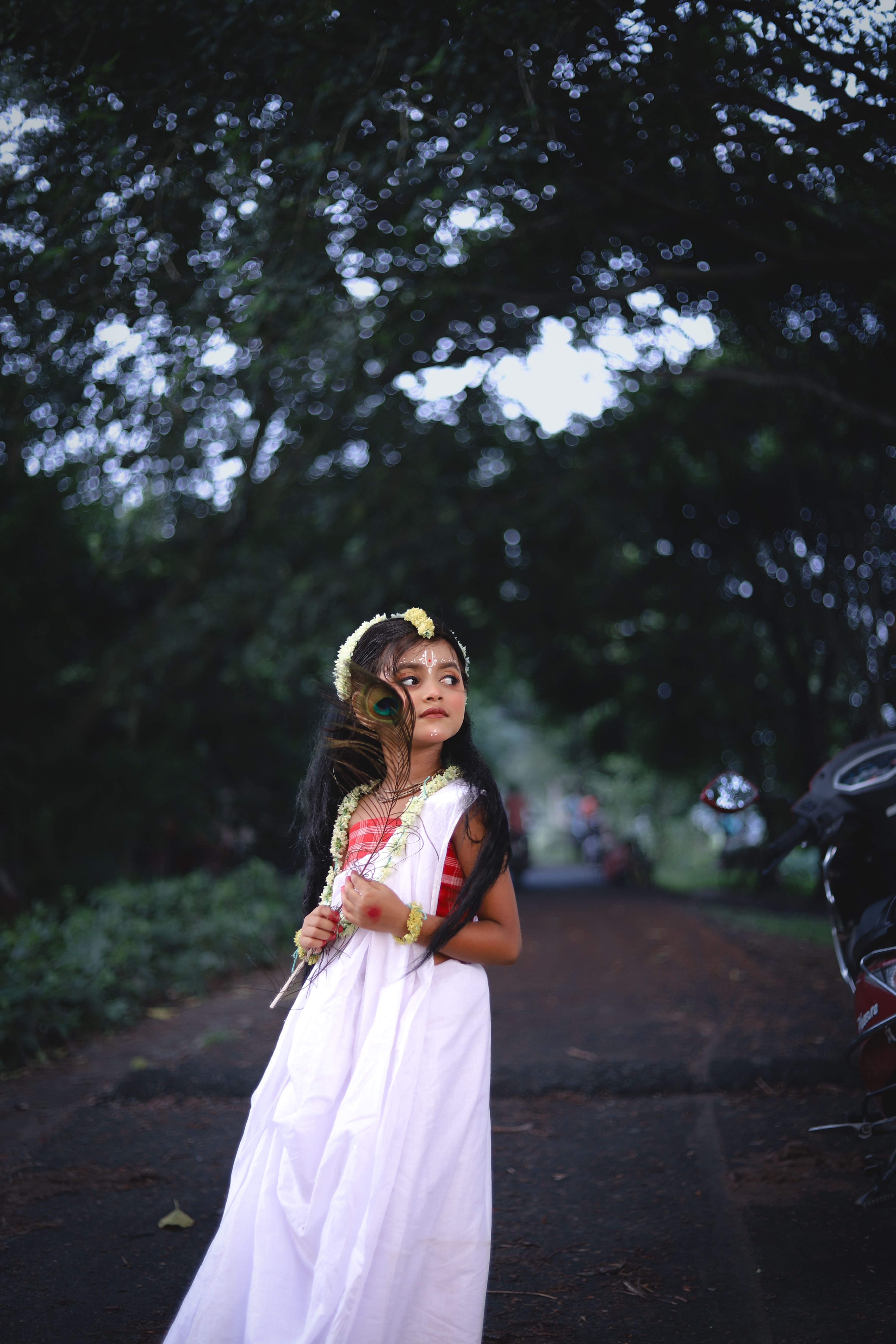 A young girl in a white dress with traditional accessories, standing outdoors in a serene setting.