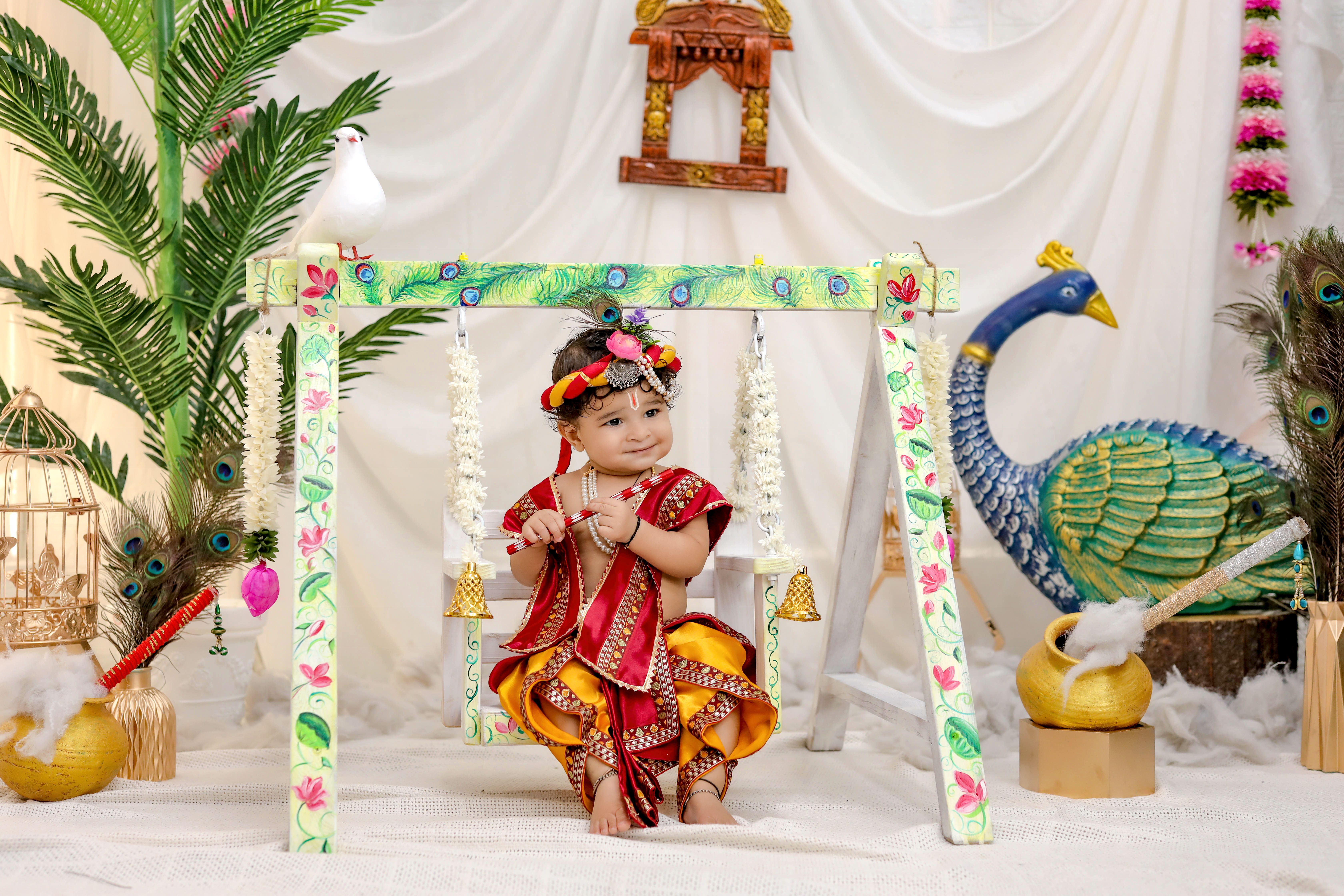 Cute baby dressed in traditional Indian clothing sitting on a decorated swing indoors.