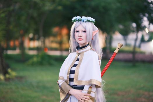 A cosplayer dressed as an elf with long silver hair poses outdoors in a tranquil park.