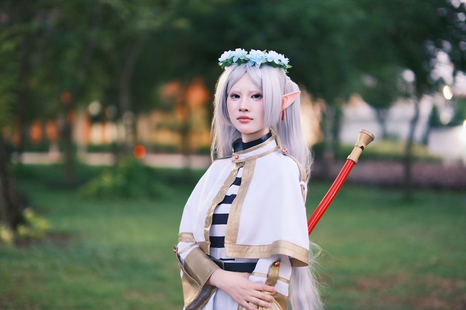 A cosplayer dressed as an elf with long silver hair poses outdoors in a tranquil park.