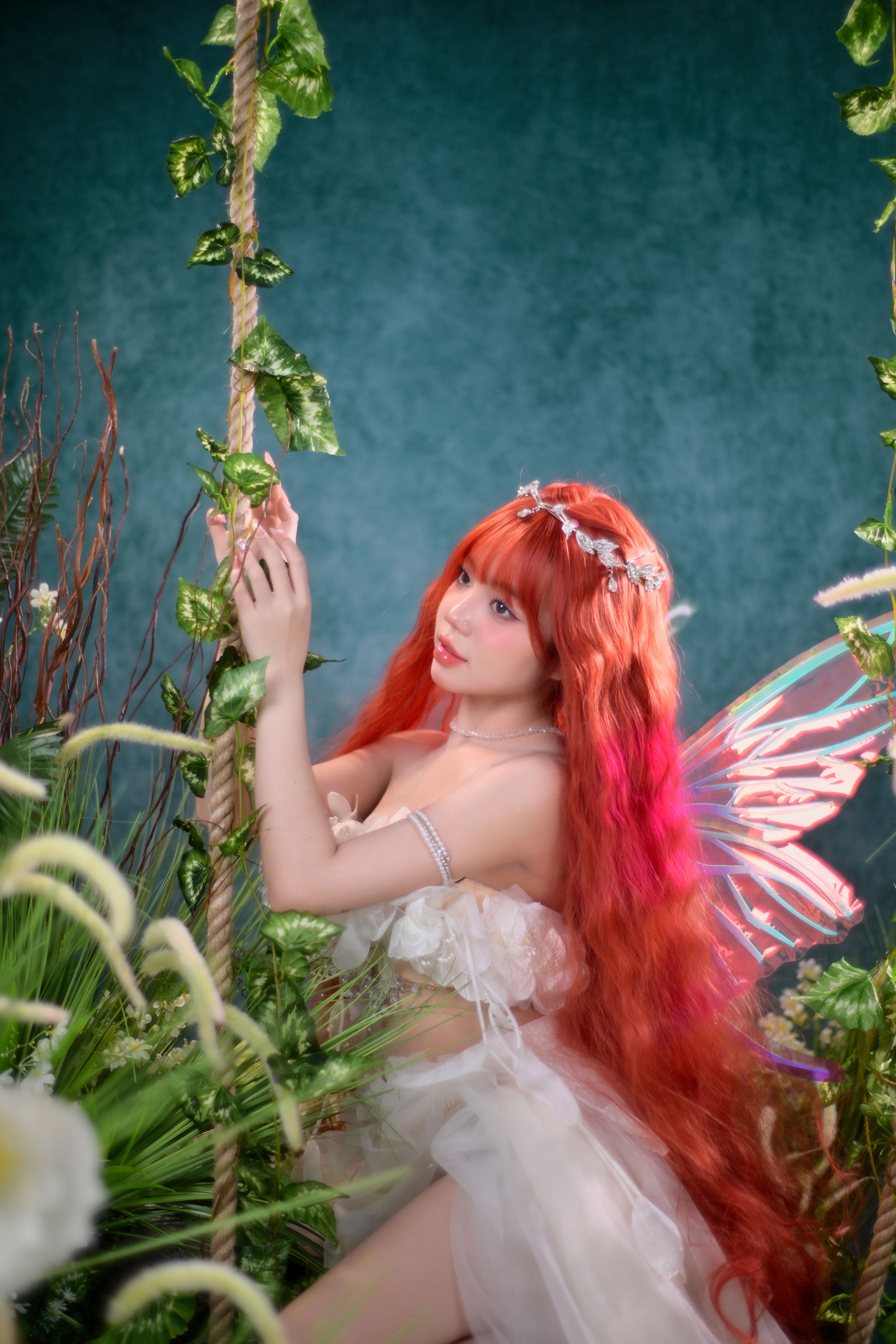 A fairy with vibrant red hair, wearing a floral crown and wings, in a lush green forest setting.