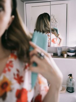 A woman in a floral dress brushes her hair in front of a mirror in a bathroom.