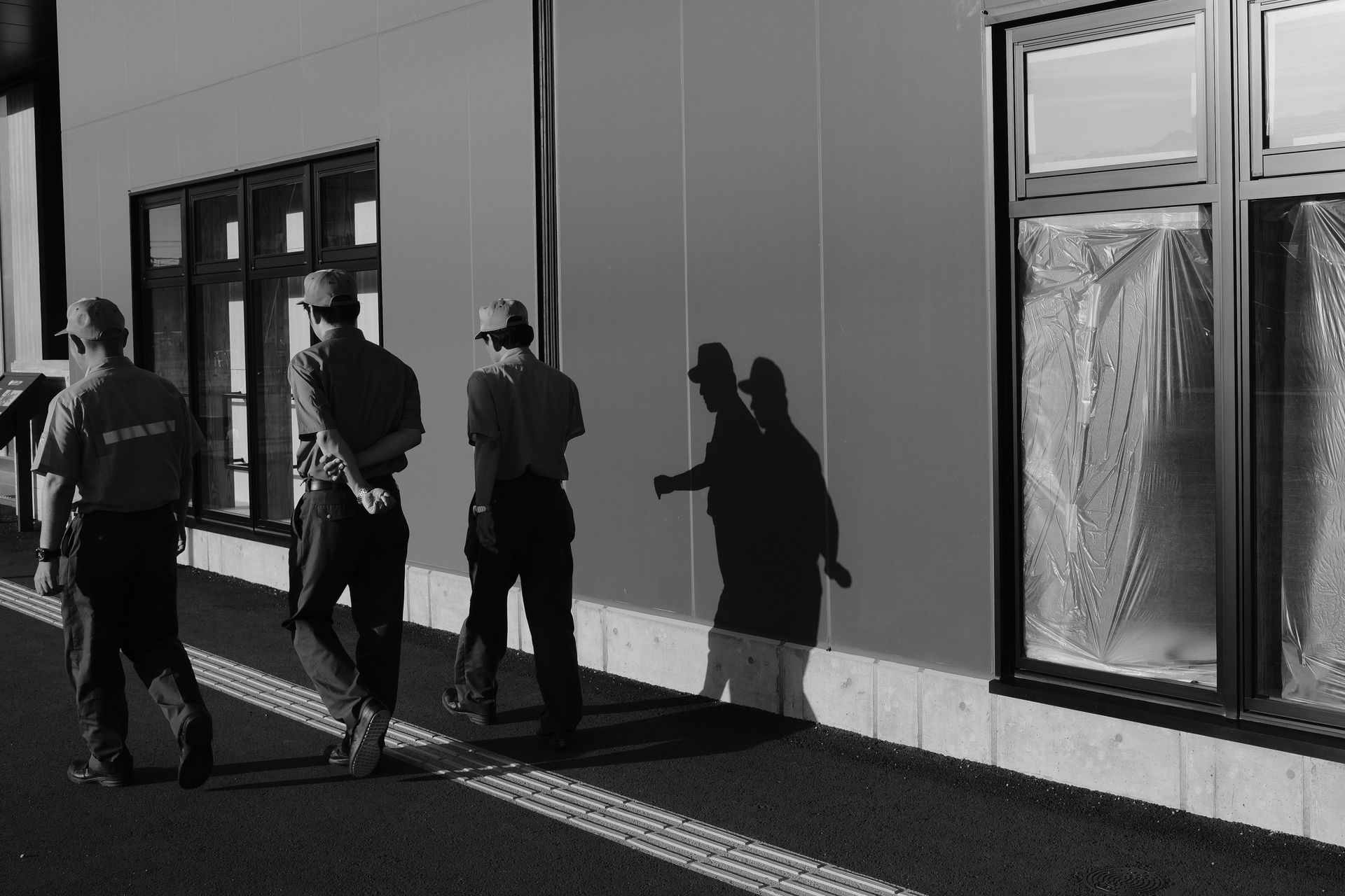 Black and white photo of workers walking along a building casting prominent shadows.