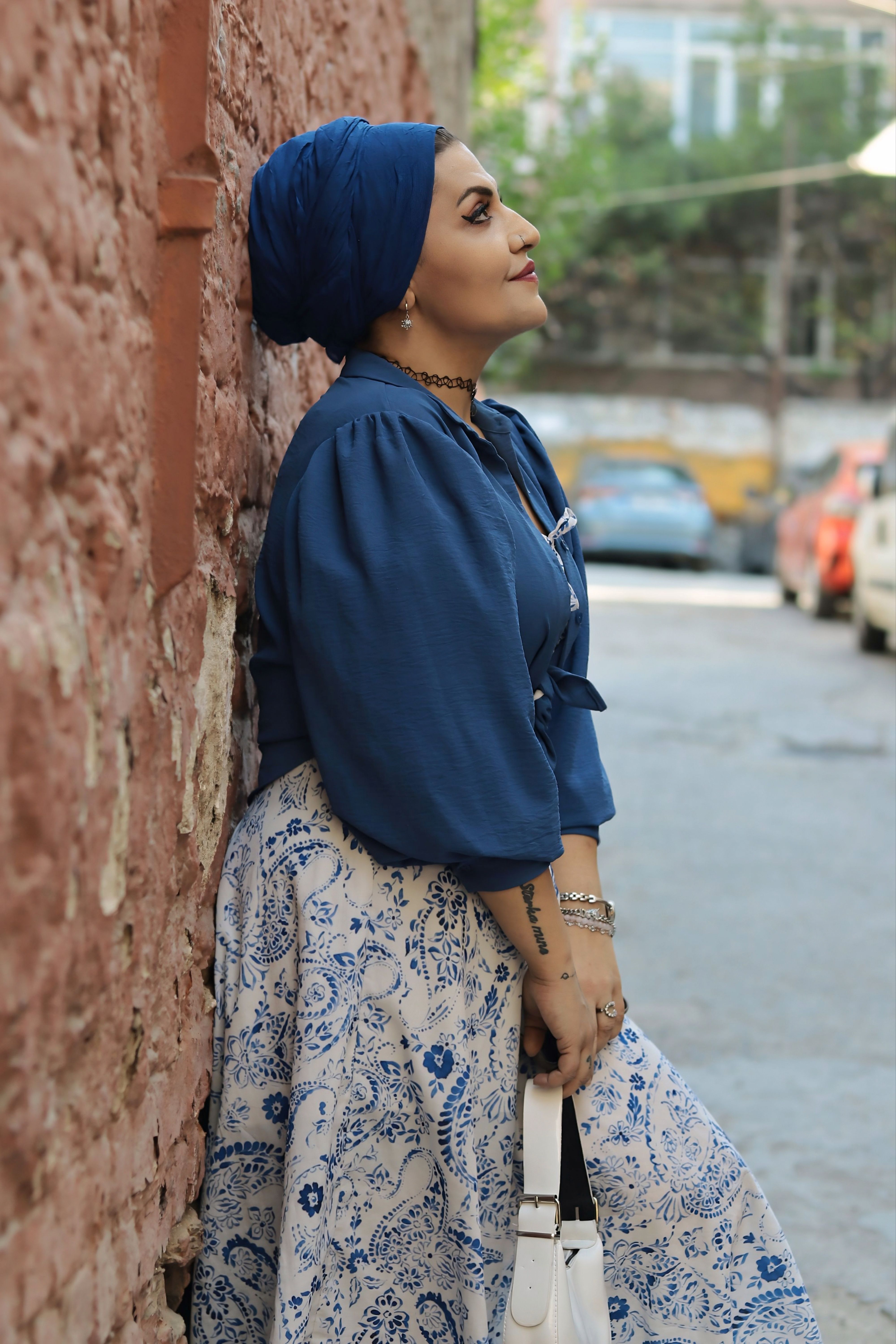 Stylish woman in blue attire leaning on a brick wall, urban backdrop.
