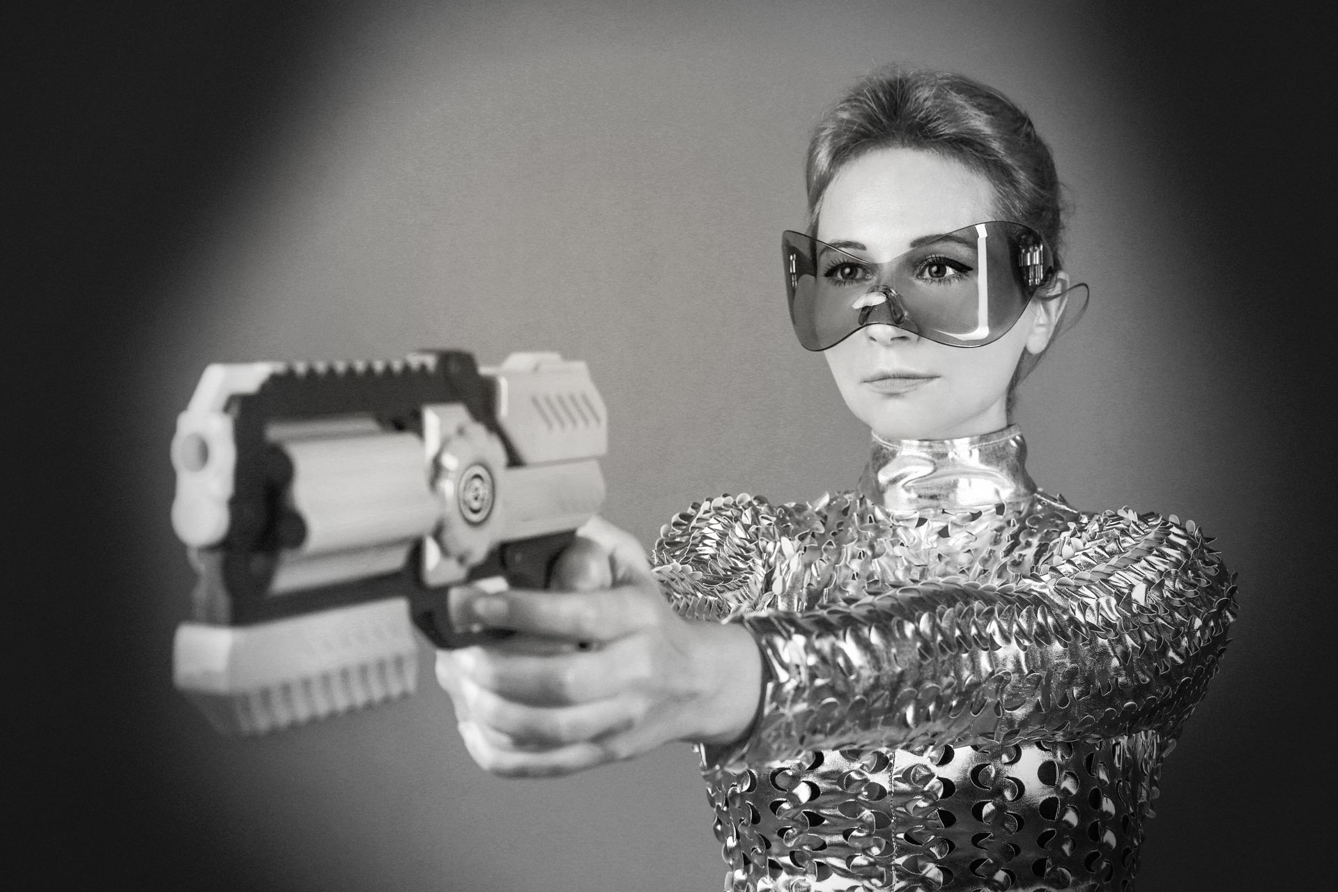 Woman in futuristic attire aiming a toy gun, wearing oversized glasses. Black and white.