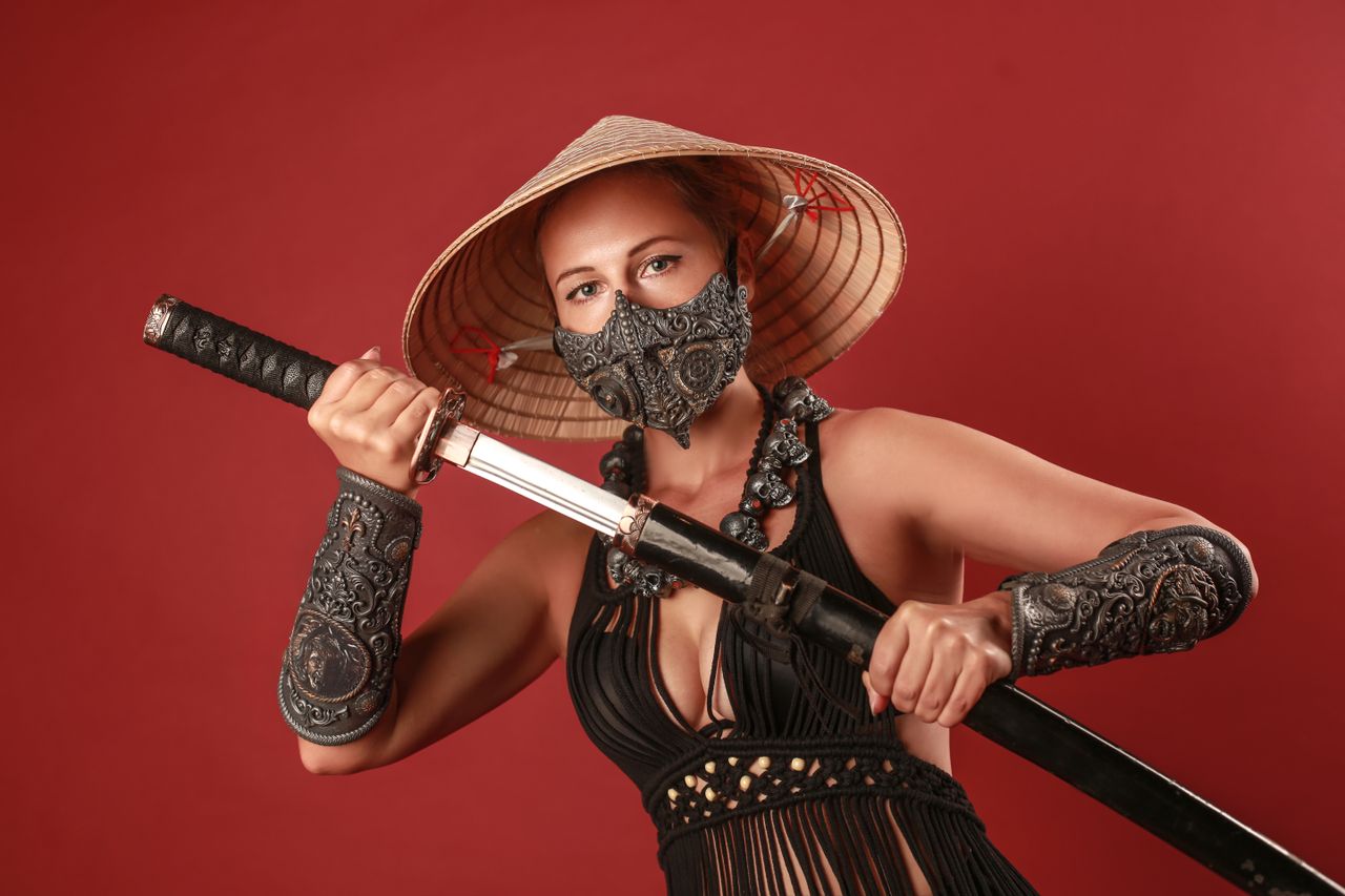a woman in asian inspired armor holds a katana against a red backdrop
