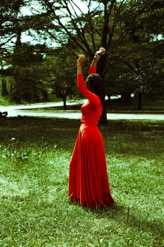 A woman in a vibrant red dress dancing gracefully in a lush green park, arms raised triumphantly.