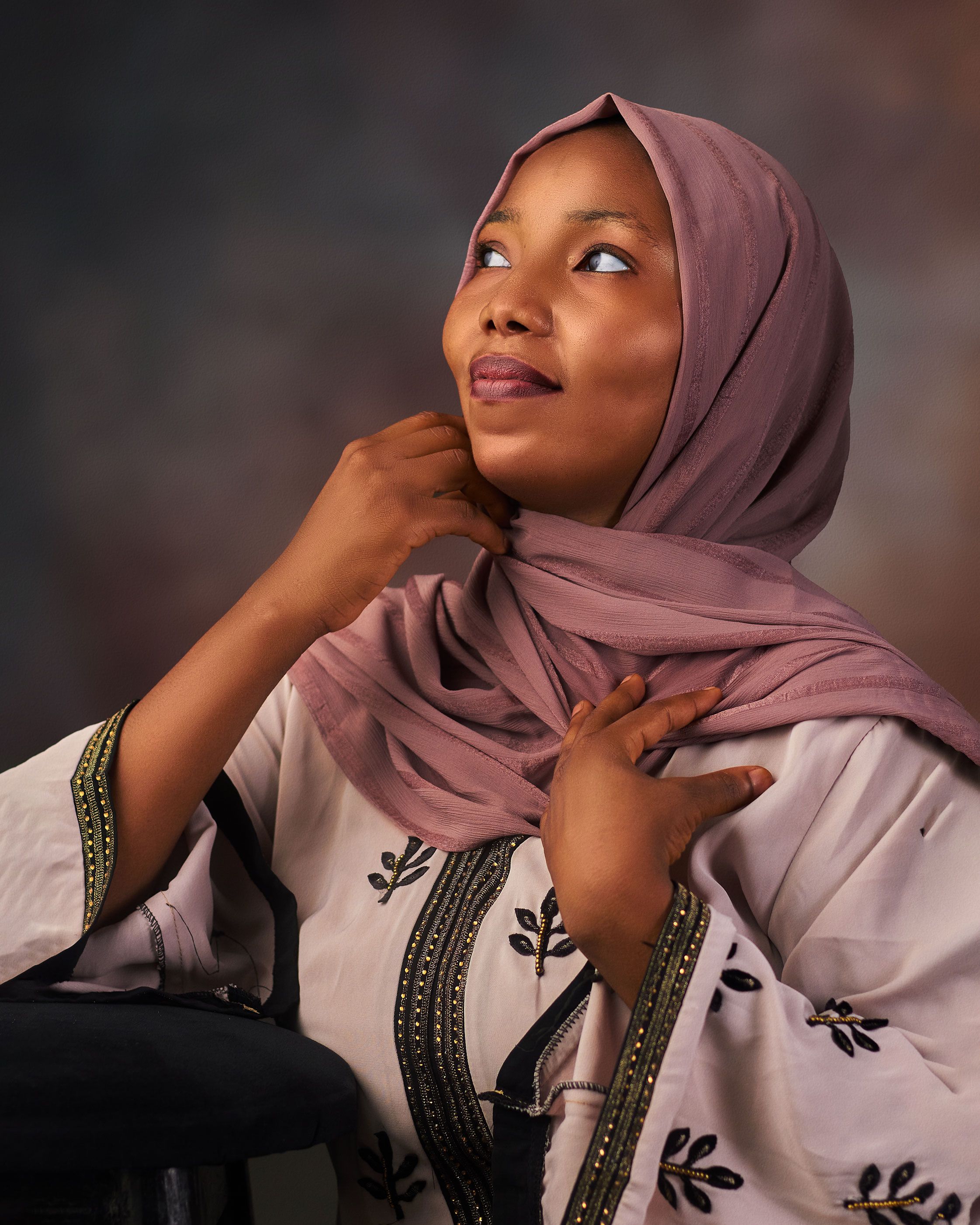A thoughtful woman wearing a pink hijab and embroidered dress, looking upwards with a serene expression.