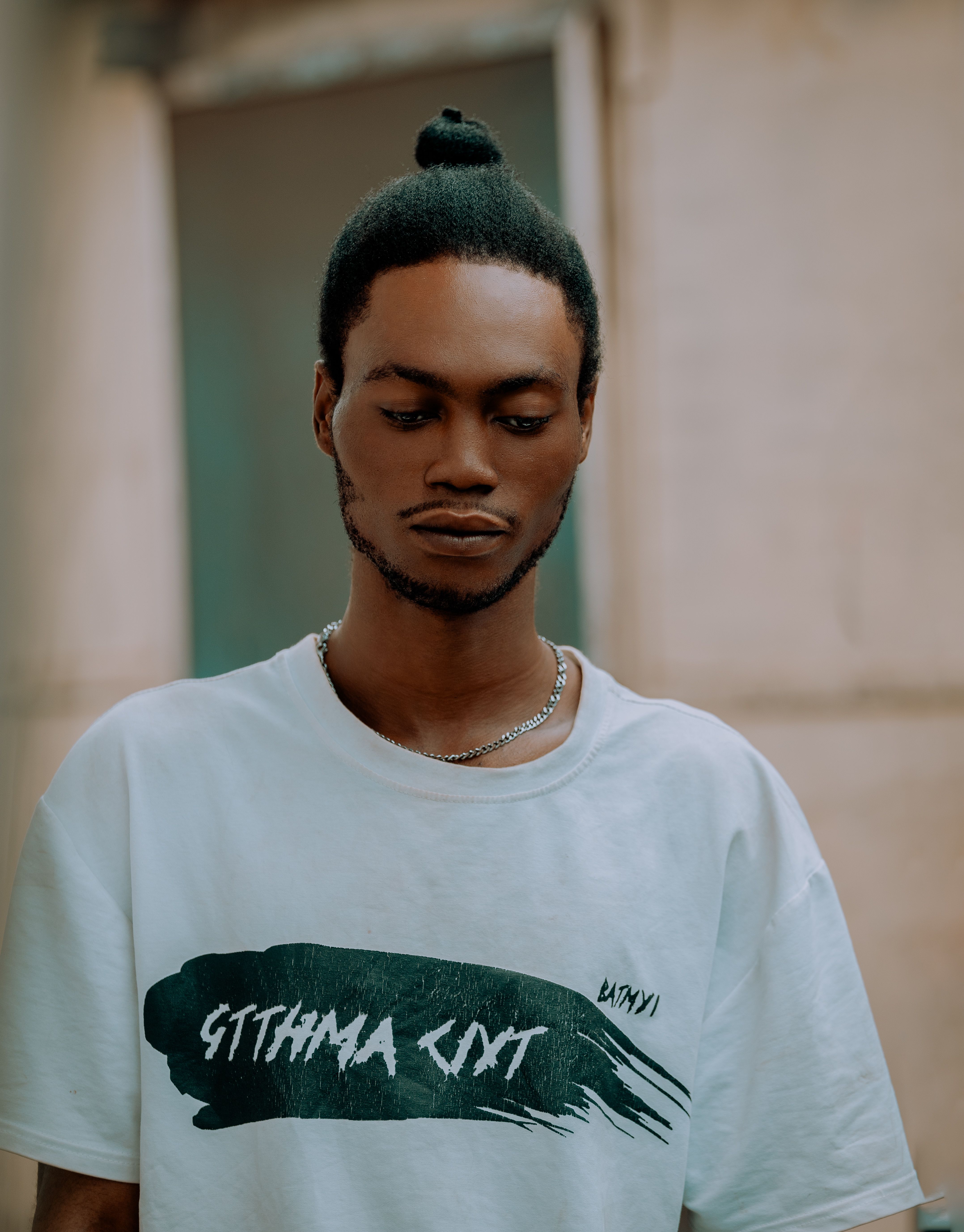 A young man with a top knot poses thoughtfully against a blurred outdoor background.