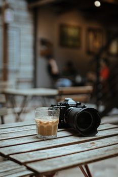 A camera and iced coffee rest on a rustic table in an atmospheric cafe setting.