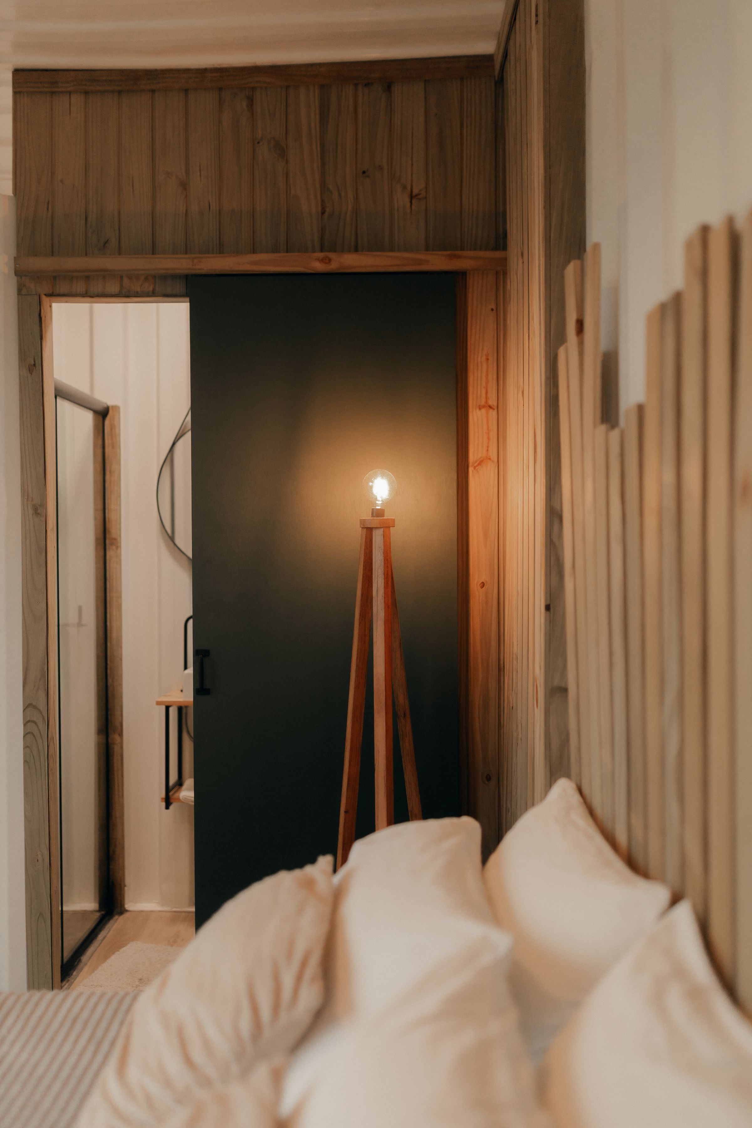 Warm bedroom with vertical timber paneling, a softly lit bed, brown armchair and glowing tripod lamp