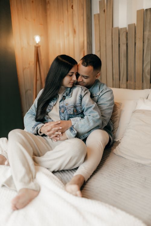 Free A couple sitting together on a bed in a cozy, warmly lit room wearing casual denim and loungewear. Stock Photo