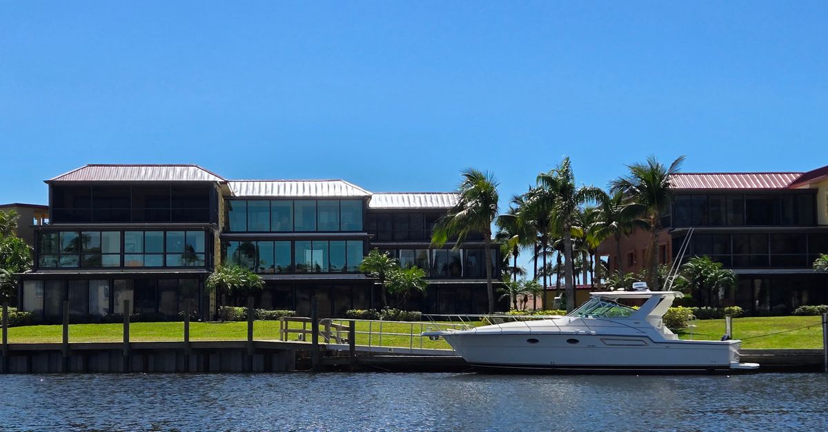 A luxurious waterfront villa accompanied by a yacht and palm trees under a clear blue sky.